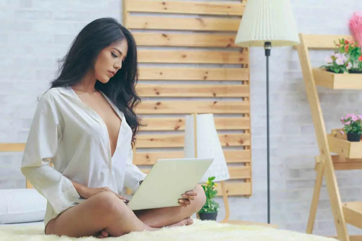 Femme brune en chemise blanche assise en tailleur sur un lit, un ordinateur portable ouvert sur les genoux