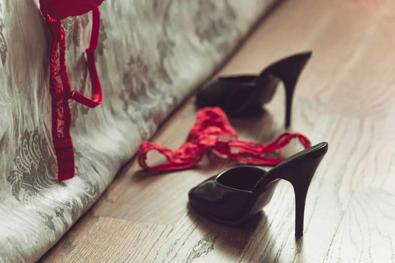 Escarpins noirs, string et soutien-gorge en dentelle rouge abandonnés sur un parquet au pied d'un lit