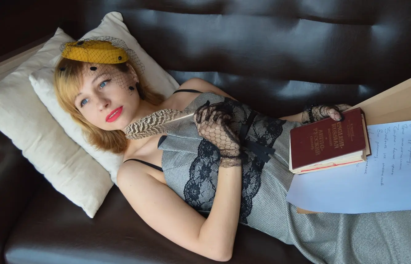 Femme blonde en nuisette et gants de dentelle allongée sur un canapé, un livre relié et des feuilles à la main