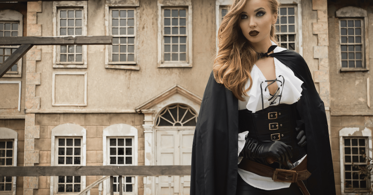 Femme blonde en cape noire et corset lacé devant la façade en pierre d'un manoir